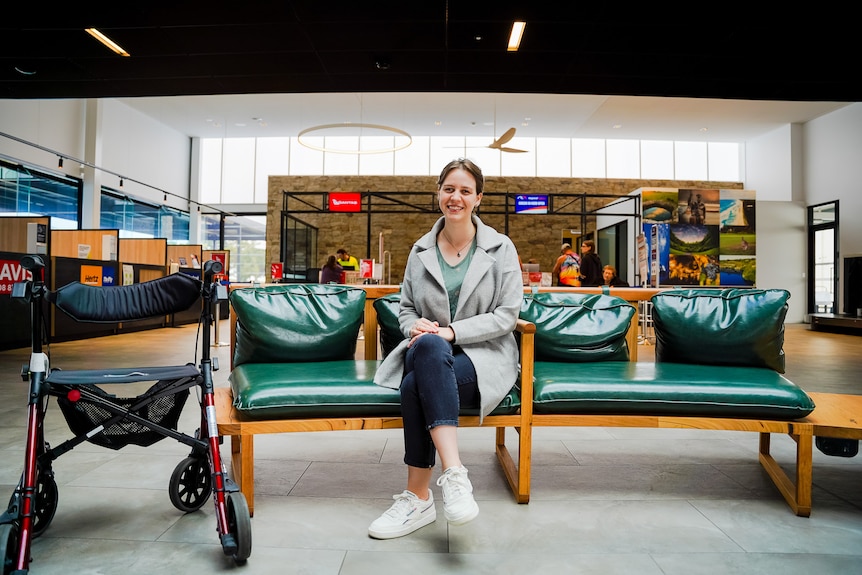Молодая женщина сидит на длинном зеленом стуле в терминале аэропорта, рядом с ней припаркована ходунка.