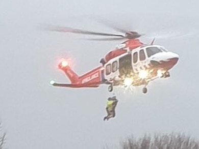 Two people hang on a cable below a helicopter hovering above floodwaters.