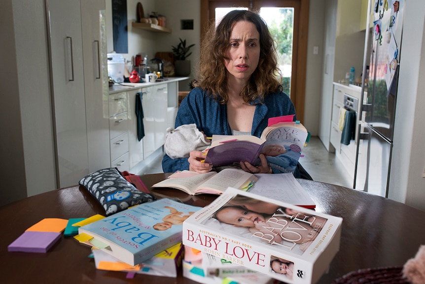 A woman with worried expression sits at a kitchen table stacked with baby books, she is also holding a baby and open book.
