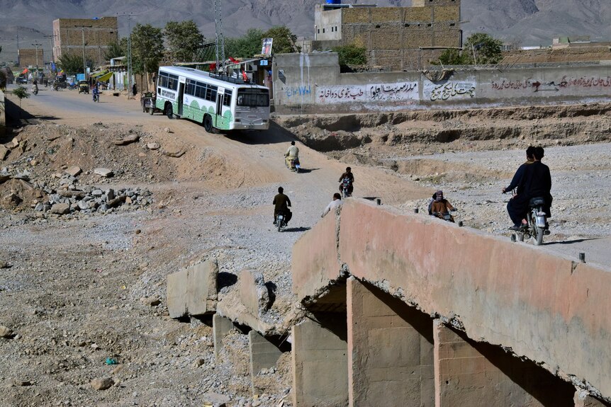 A bus and several motorcyclists navigate a damaged stone and dirt bridge which now dips close to the ground.