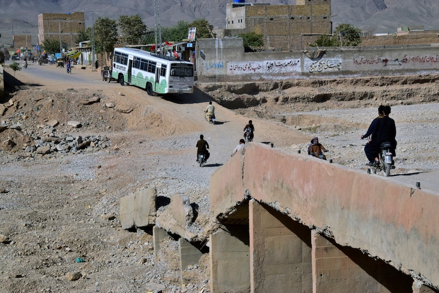 A bus and several motorcyclists navigate a damaged stone and dirt bridge which now dips close to the ground.