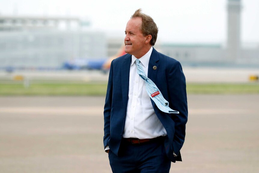 A man in a navy suit stands on the tarmac of an airport with his hands in his pockets.