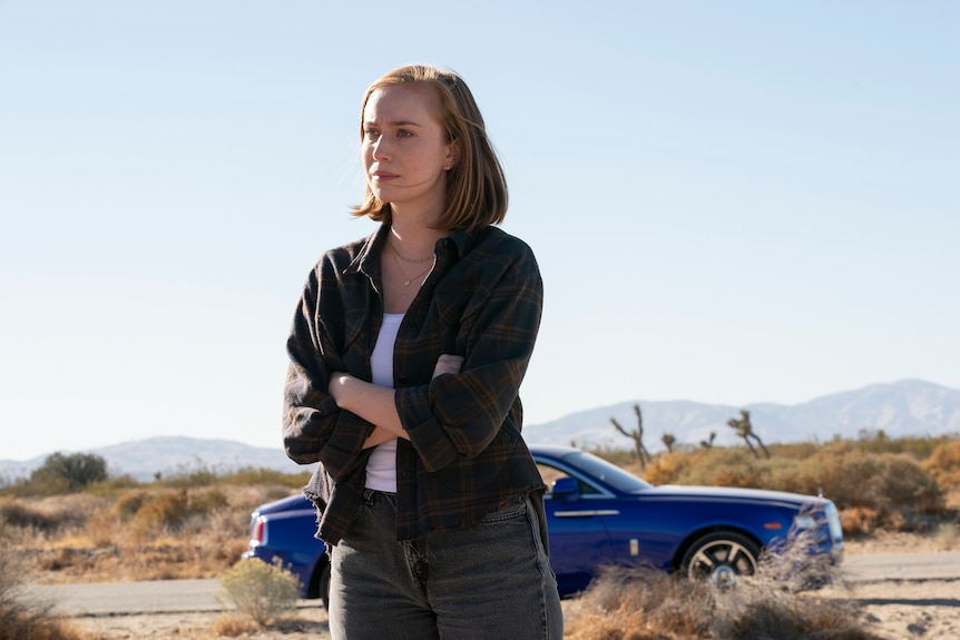 A 25 year old woman with red hair, wearing a white tshirt and dark jacket, stands in a desert with a blue car behind her