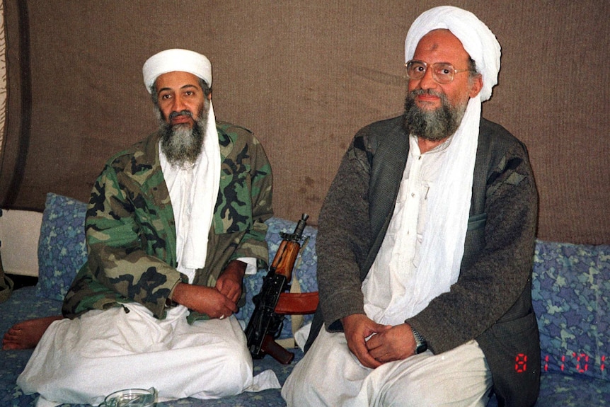 Osama bin Laden and Ayman al-Zawahiri in army fatigues sitting on the floor next to a rifle