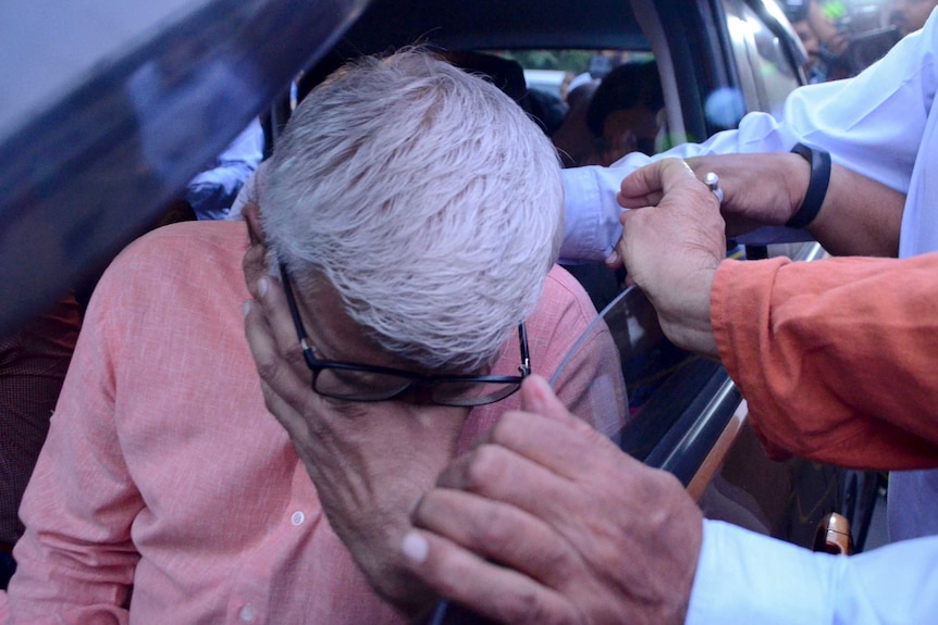 A man is seen with his head in his hands while sitting in a car with people surrounding it.