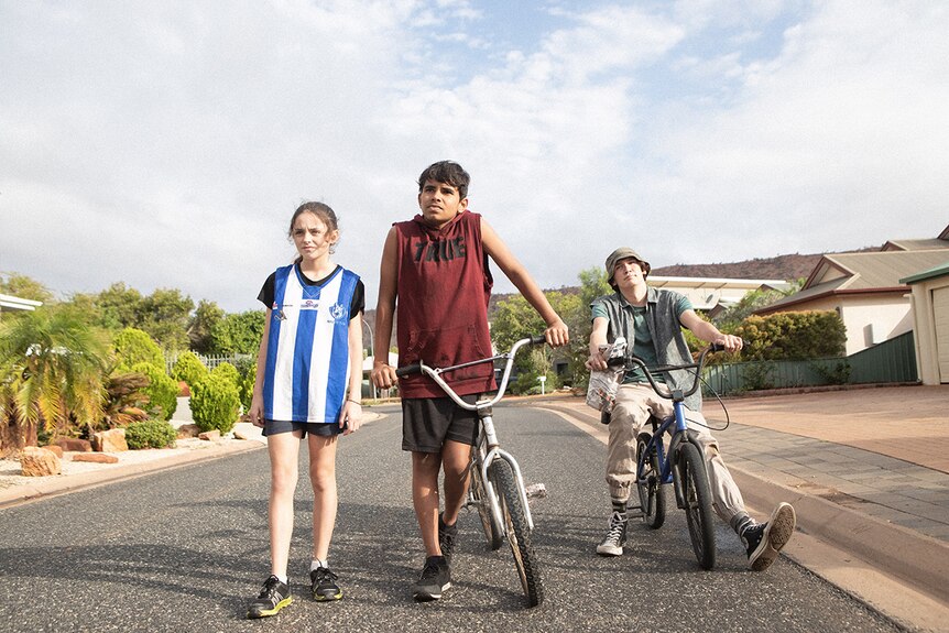 Teenage girl wearing striped AFL jersey and two teenage boys with BMX bikes walk down suburban street on a bright cloudy day.
