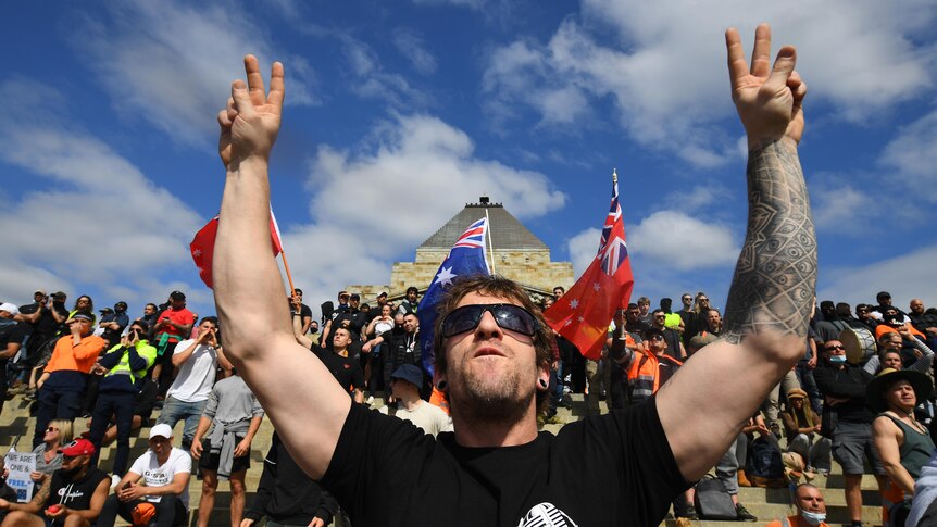 A man with sleeve tattoo on one of his arm makes a victory sign with lots of people sitting behind him and flag of Australia 