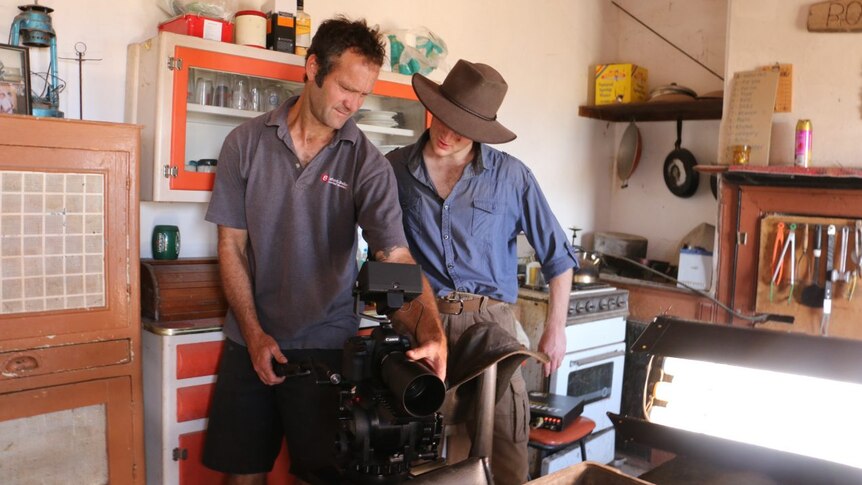 A man in a grey shirt and a man in a blue shirt stare into the back of film camera and discuss the footage.