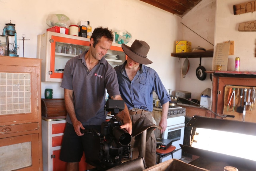 A man in a grey shirt and a man in a blue shirt stare into the back of film camera and discuss the footage.