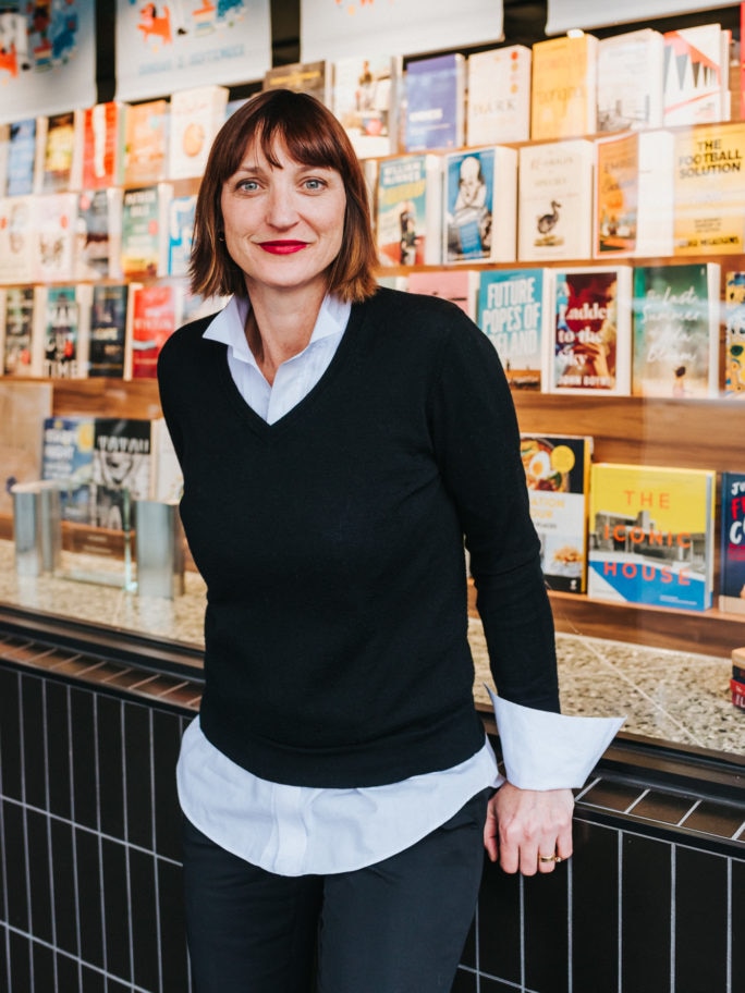 A 40-something brunette woman with a bob haircut leans back against a counter in a bookshop, books lining the wall behind her