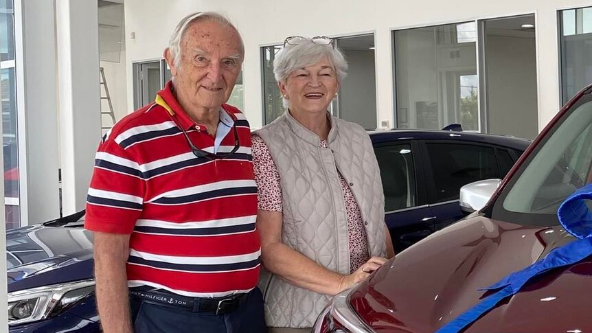 Retirees, Pat and Lyndon Rae stand together next to a car with a blue ribbon