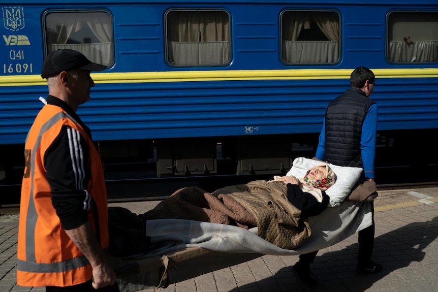 An old woman lying on a stretcher is carried towards a train.