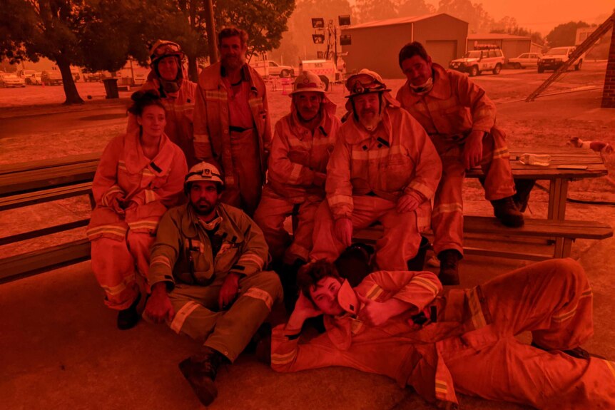 A group of firefighters pose for a photo, the sky and photo is very orange from the smoke haze.