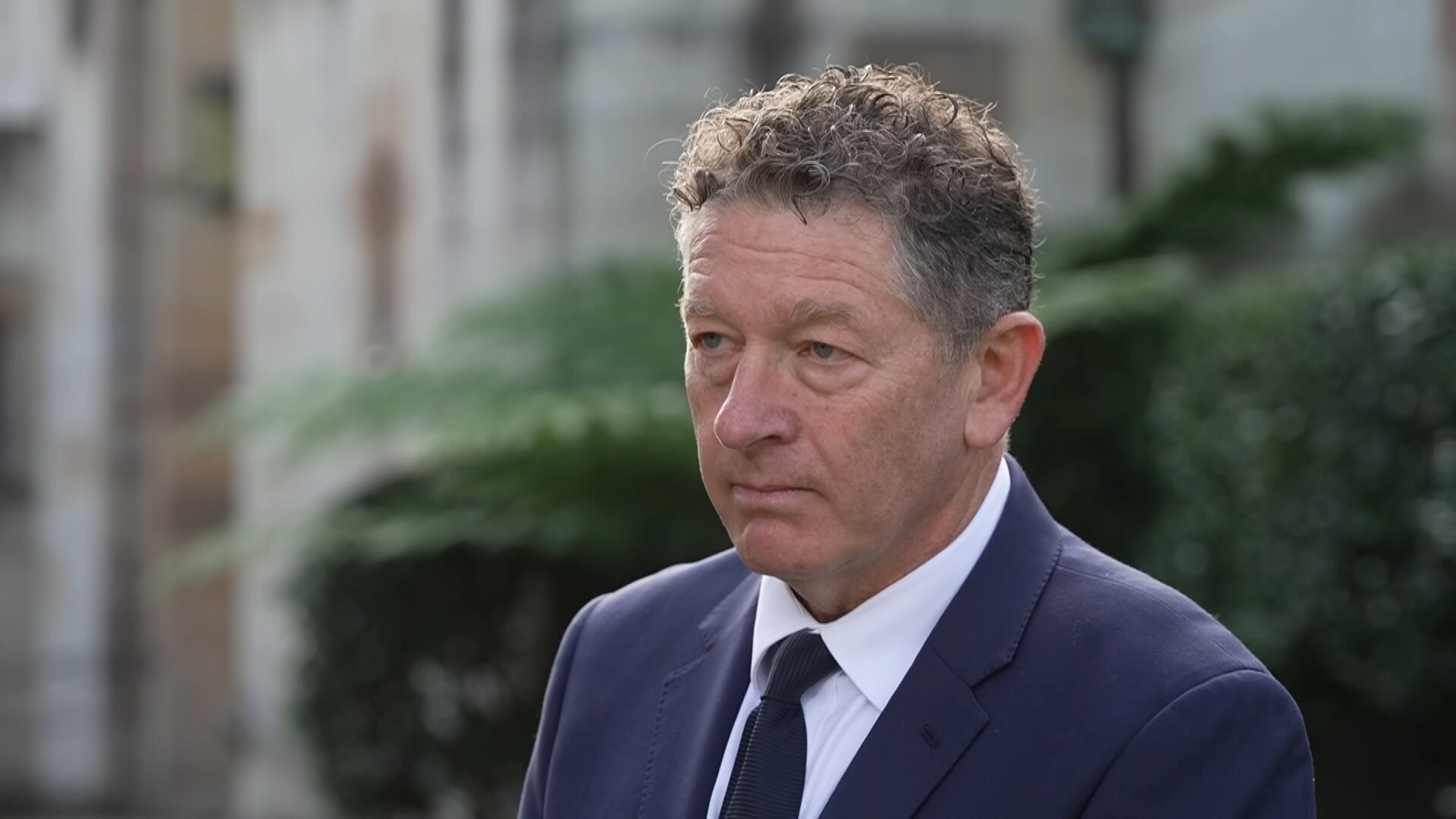 A middleaged man wearing a navy suit and tie, stands solemnly outside a church.