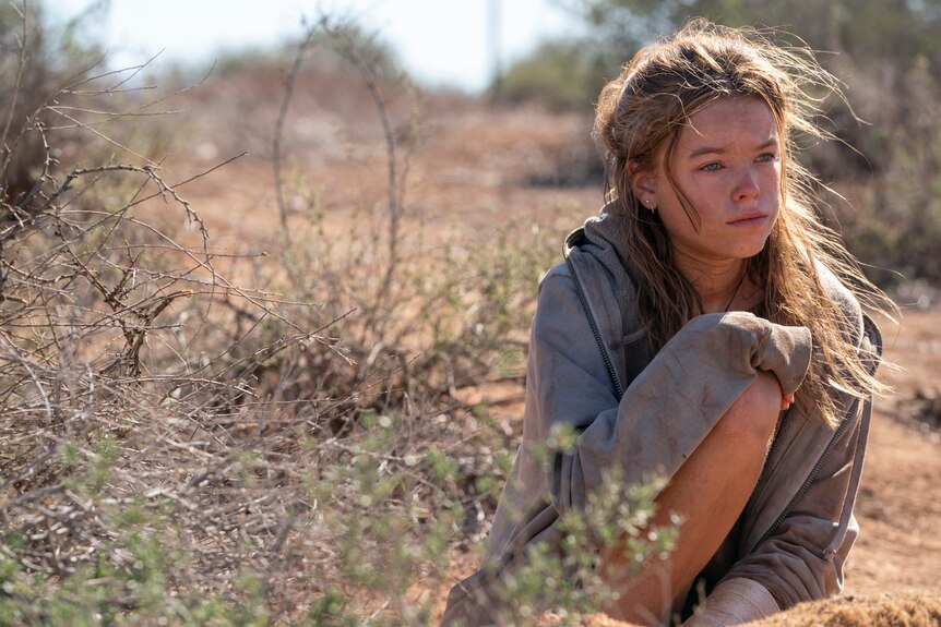 A long-haired teenage girl, covered in dirt, crouches in the desert, looking morose