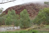The red moutain range is shrouded in grey low hanging cloud above the brown flowing Todd River.