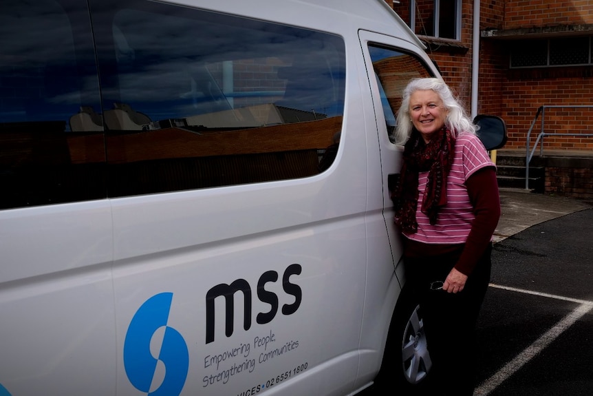 A woman in her 60s is standing beside the driver door of a mini bus with MSS logo on the side.