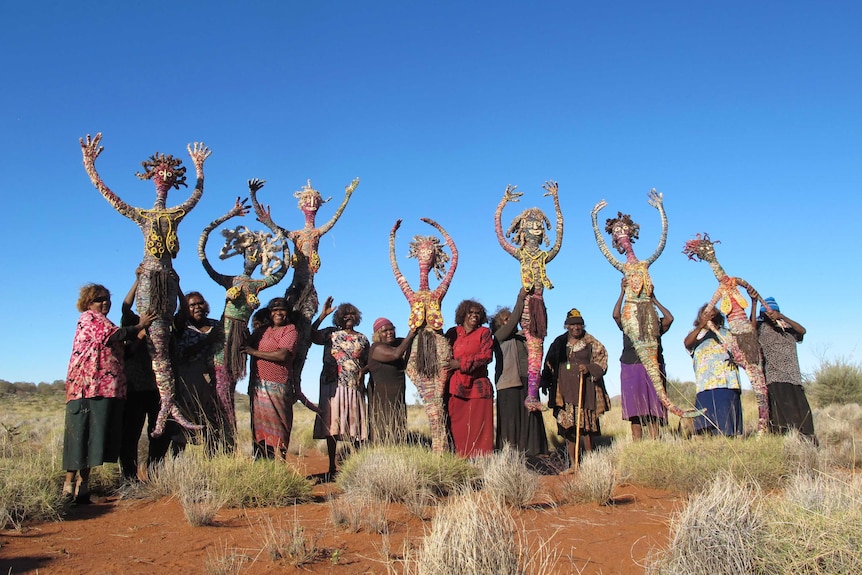 Eleven women, the Tjampi Desert Weavers, stand with their artworks, the Seven Sisters figures.