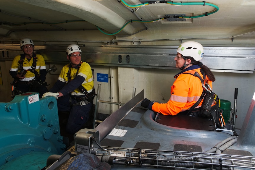 Three people in high vis and helmets stand inside a small engine room.