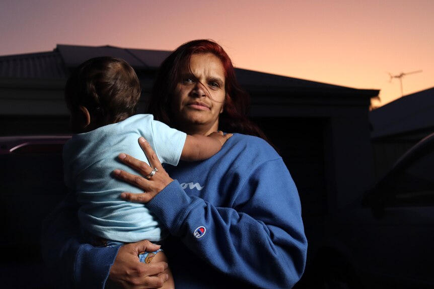 A woman stands holding a toddler in front of a house with a neutral expression. The sky is orange from the sunset. 