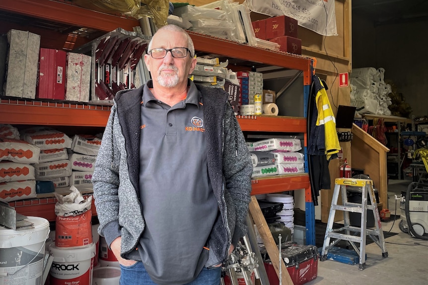 A man stands in a shed with plastering equipment behind him