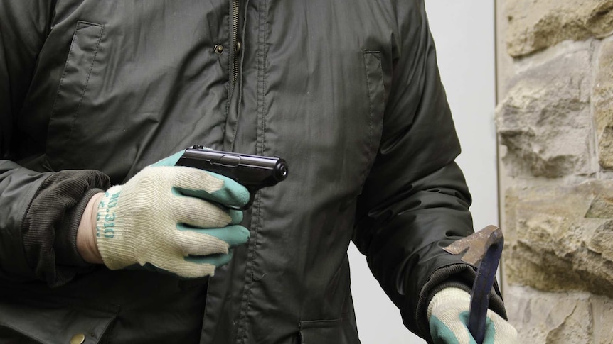 A burglar in a black jacket and wearing gardening gloves holds a pistol and crowbar.