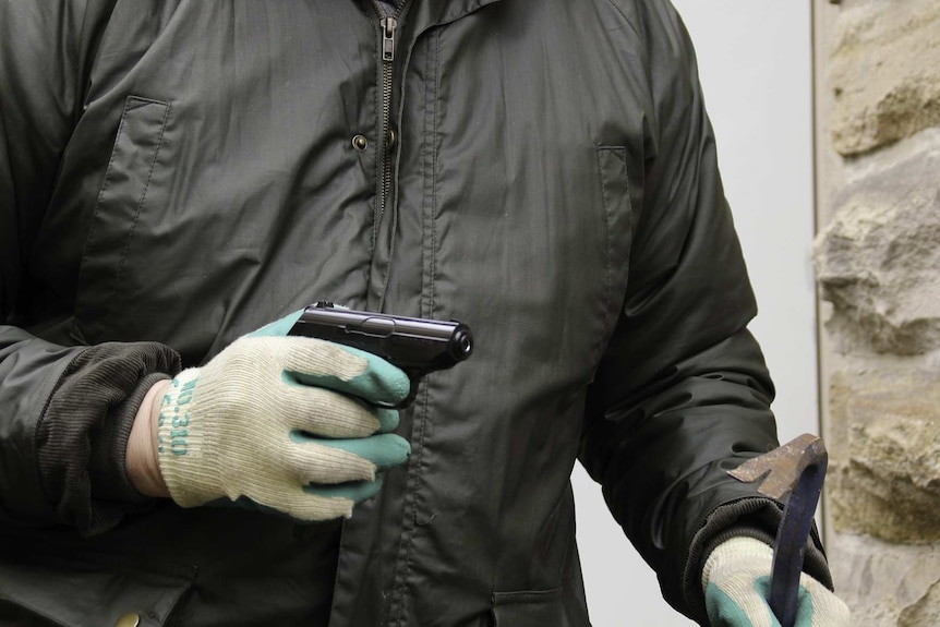 A burglar in a black jacket and wearing gardening gloves holds a pistol and crowbar.