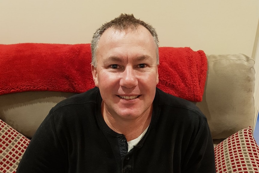 A white man, wearing a black long sleeved tshirt looks at a camera and smiles, while sitting on a couch