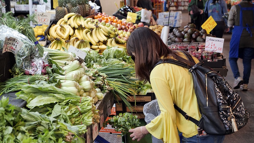 A woman wearing a yellow cardigan browses green leafy vegetables at an indoor market