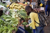 A woman wearing a yellow cardigan browses green leafy vegetables at an indoor market