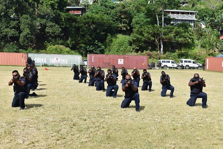 Training police kneel on grass and point their guns towards the camera.