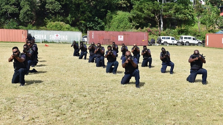 Training police kneel on grass and point their guns towards the camera.