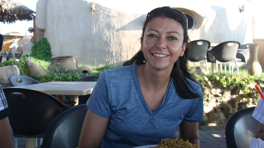 A medium shot of a woman sitting at a table outdoors and smiling, with a plate of food in front of her.