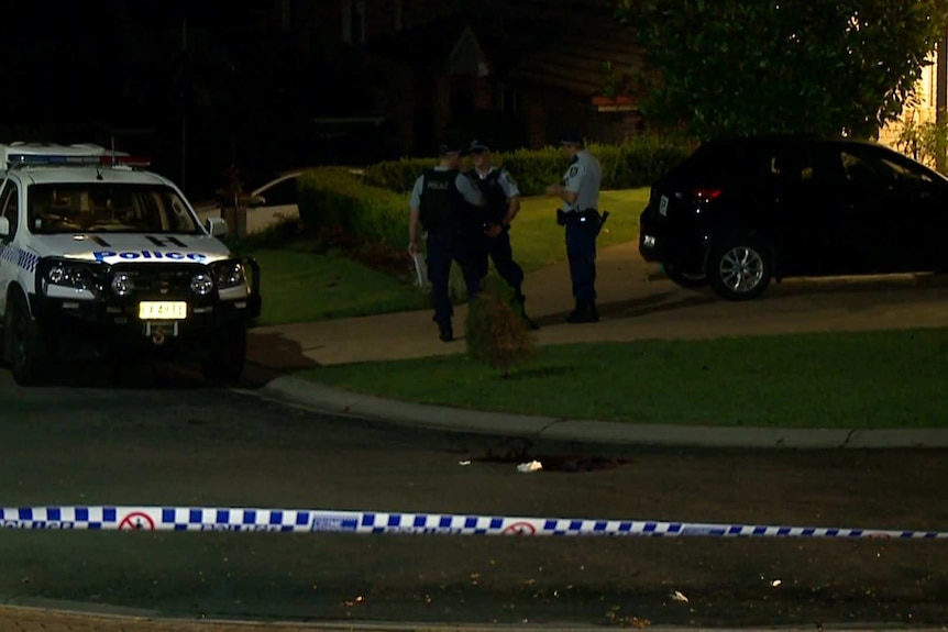 Police mill around in front of a house in a suburban street, police tape in front of them