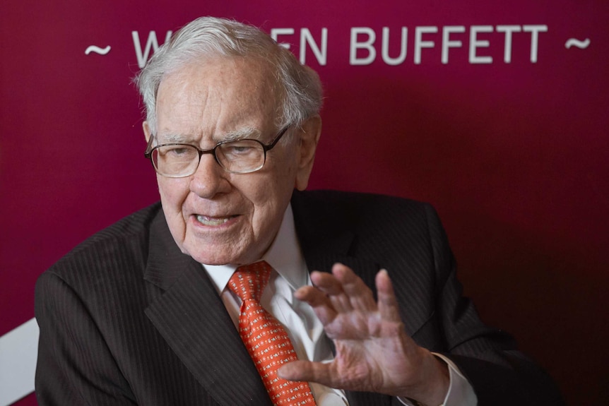 Warren Buffett, Chairman and CEO of Berkshire Hathaway, wearing a pinstripe suit and a red tie holds up his hand.