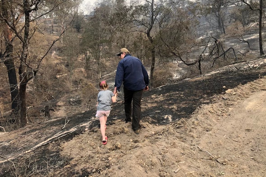 A man holds his daughter's hand as they walk through a burnt-out forest.