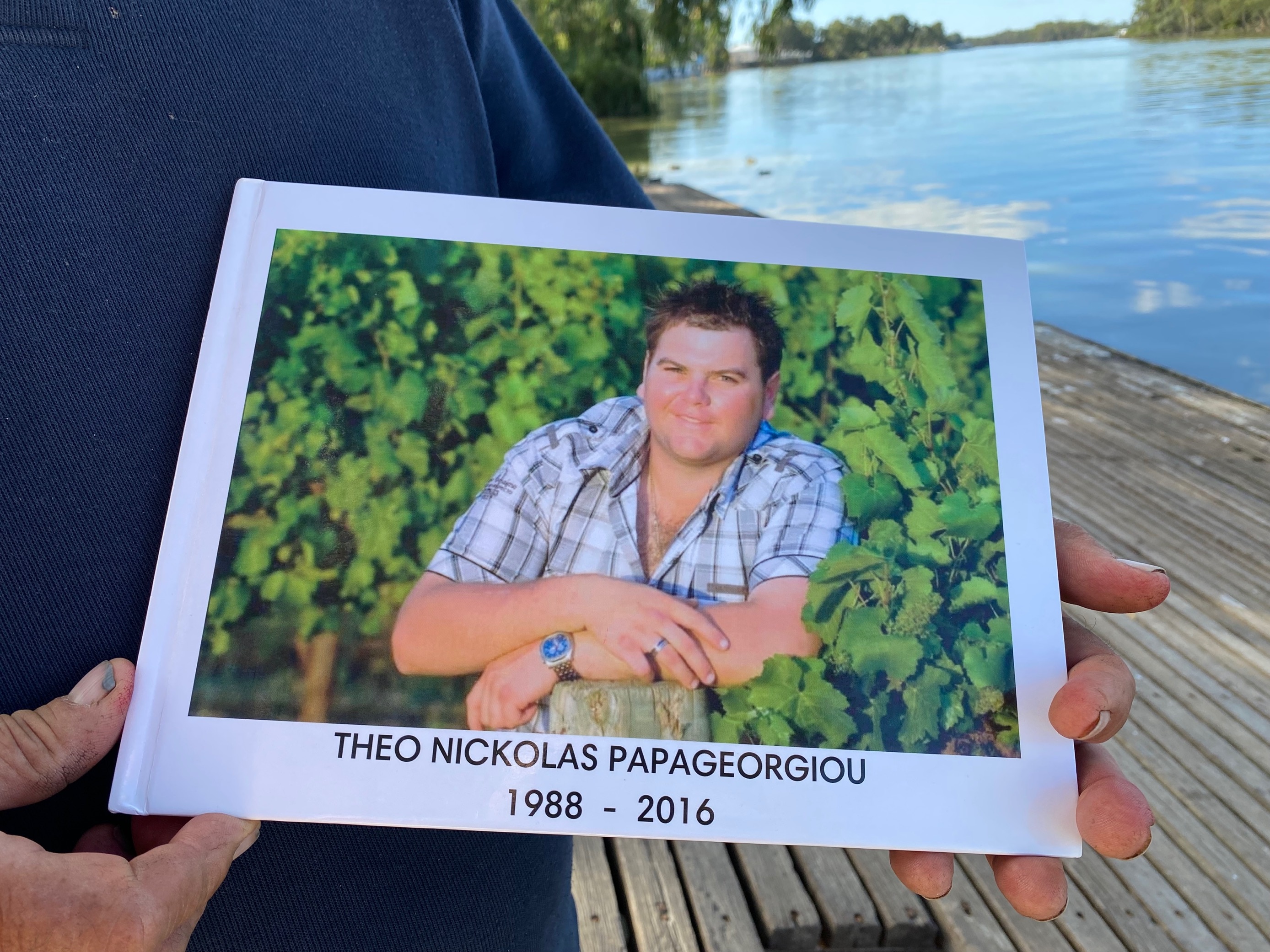 A white man with brown hair and a checked shirt stands in lush, green grape vines with a cheeky, full smile. 