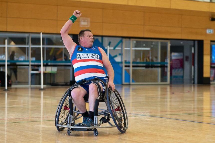 A man wearing a blue white and red guernsey sits in a wheelchair. His hand is held above his head after completing a handball