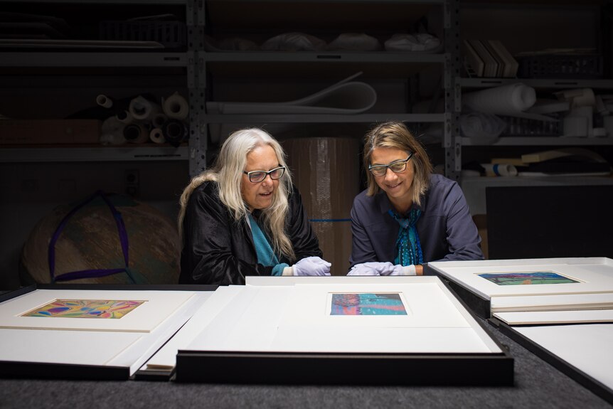 A grey-haired woman wearing glasses, sits with a brown-haired woman looking at art pieces on a table. Both smile.