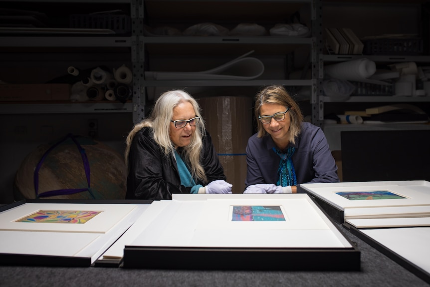 Two women sit in a dimly lit room, examining some artwork.