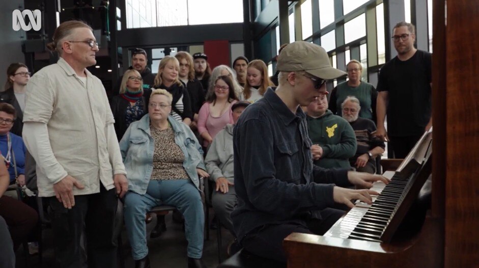Dan Duffy sitting on the piano with his back to the Find Your Voice Collective as they perform a song.