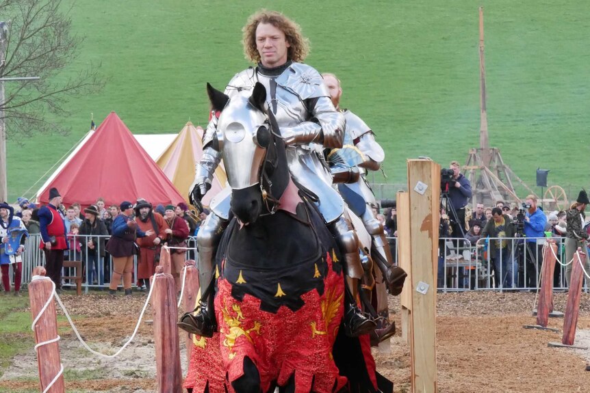 A man dressed in armour rides a horse in front of a crowd, with another man dressed as a knight behind him.
