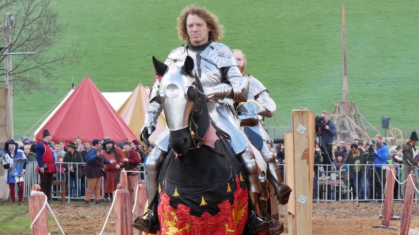 A man dressed in armour rides a horse in front of a crowd, with another man dressed as a knight behind him.