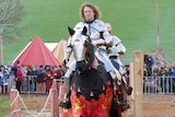 A man dressed in armour rides a horse in front of a crowd, with another man dressed as a knight behind him.