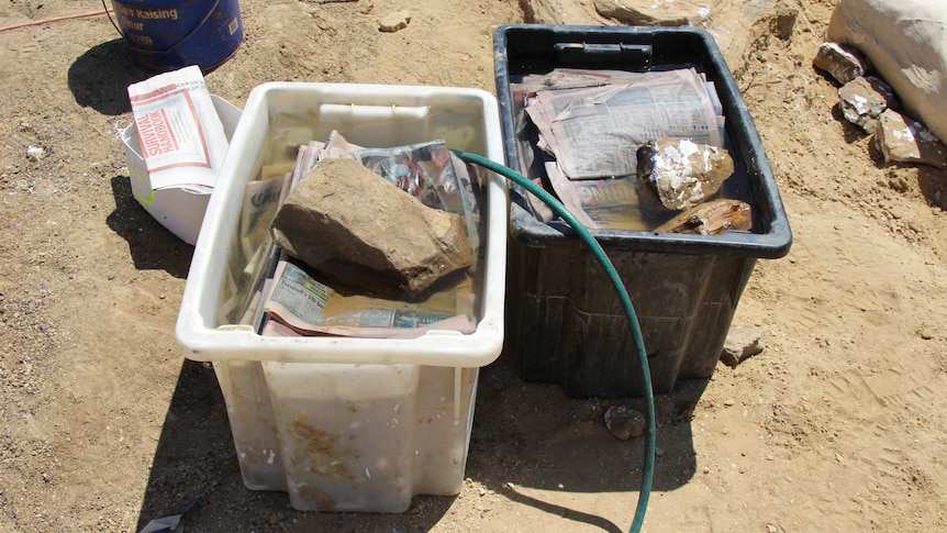 Large plastic boxes full of water, newspaper and rocks sit on the dusty ground.