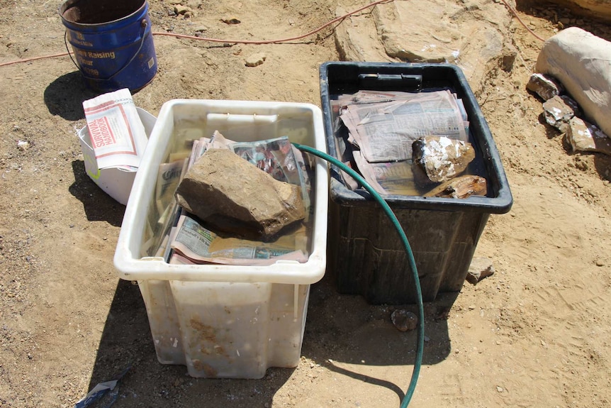 Large plastic boxes full of water, newspaper and rocks sit on the dusty ground.