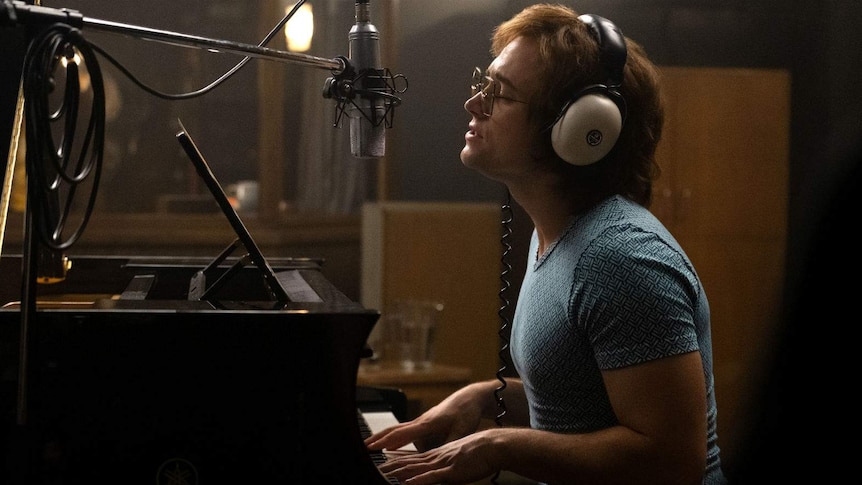The actor sits at a piano, wearing white headphones, singing emotionally into a microphone.
