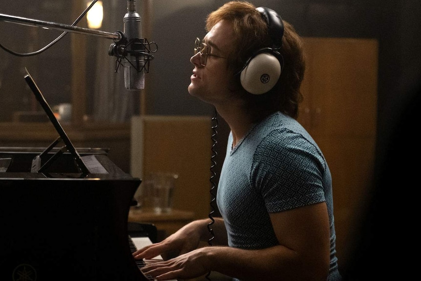 The actor sits at a piano, wearing white headphones, singing emotionally into a microphone.