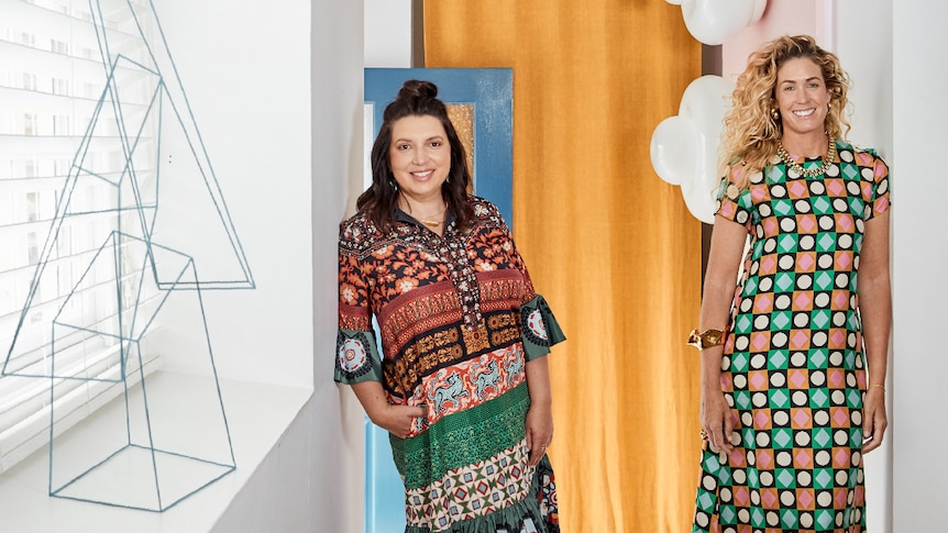 Two women in bright geometric dreses stand in a trendy decorated interior. 