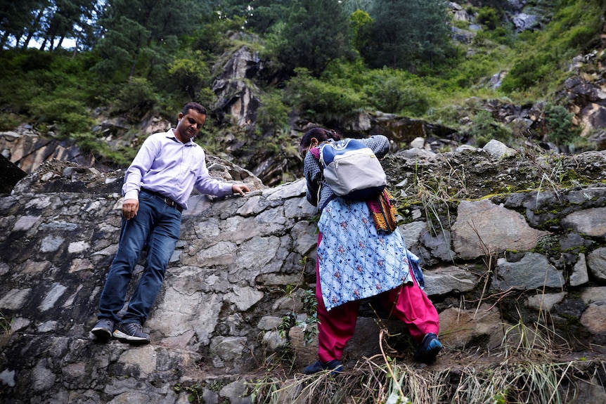 A woman grabs onto a brick wall while trekking over rocky terrain as a man watches on.